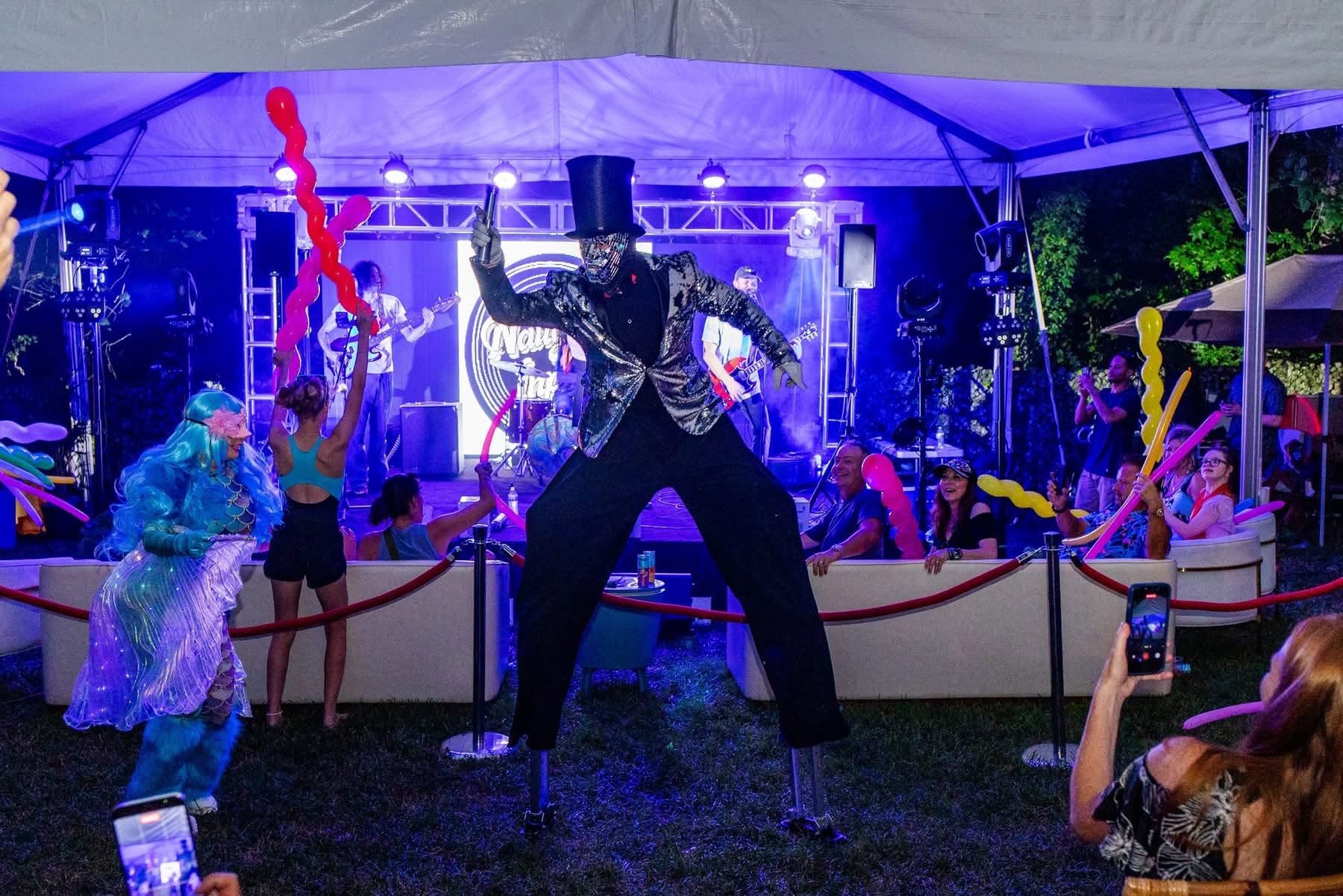 Mirror-suited stilt walker in a top hat performing under a purple-lit tent while a live band plays and guests watch with balloon swords