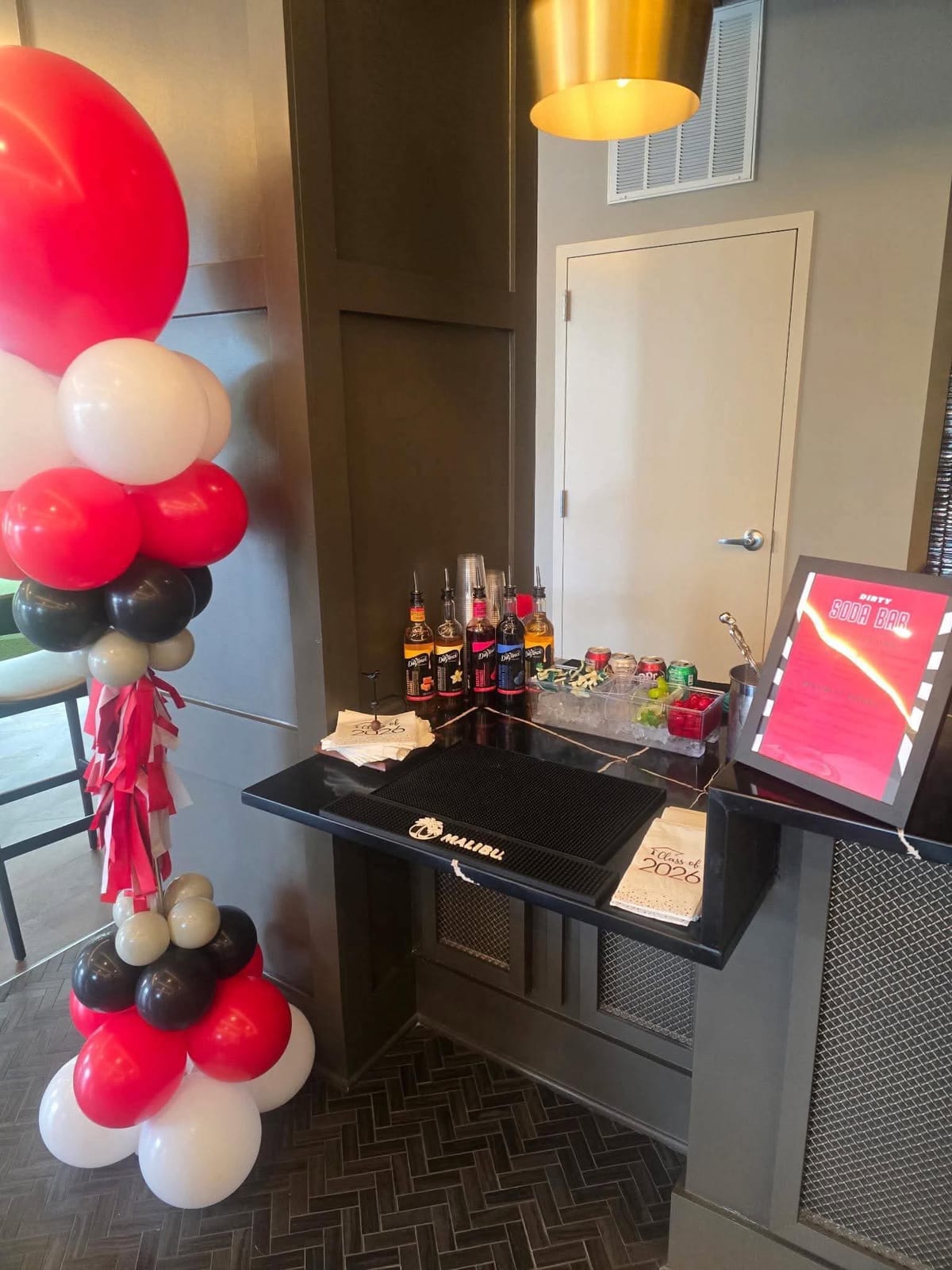 Red, black and white balloon column beside a dark bar cart with spirits and a signature drink sign