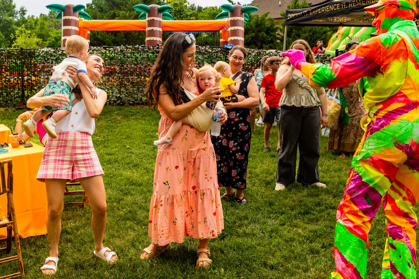 Guests holding babies as a neon-costumed performer greets them on a lawn