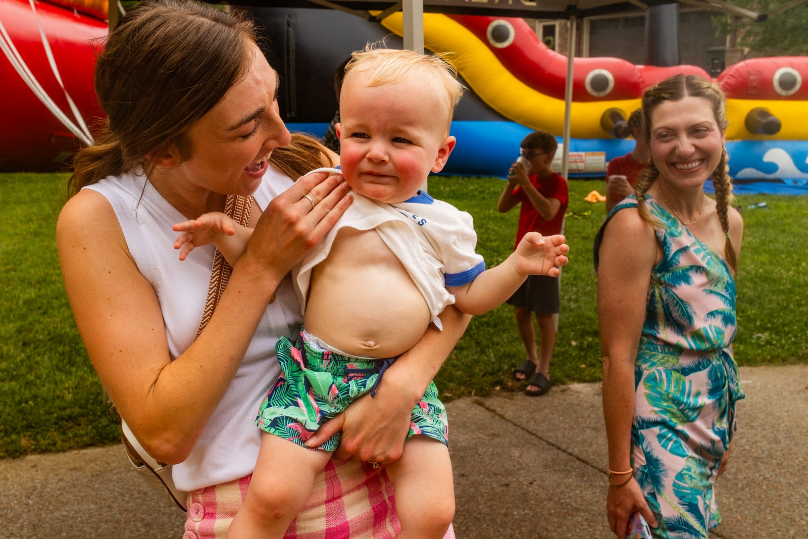 An adult feeding a baby at an outdoor party with an inflatable in the background