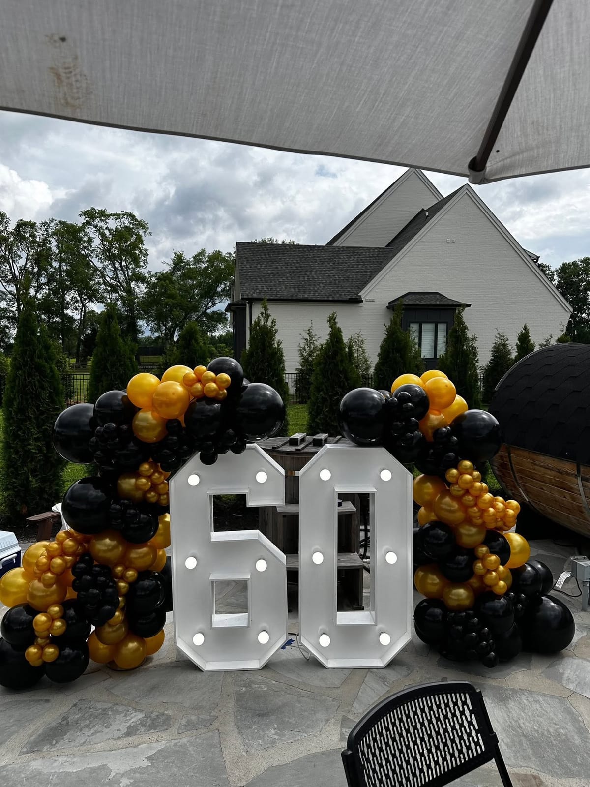 Lit marquee number sixty framed by black and gold balloon garlands outside a large home