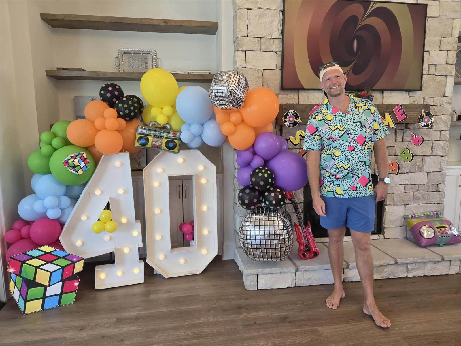 A guest in a nineties-print shirt and backwards cap beside lit marquee 40 numbers under a colorful retro balloon garland with a boom-box balloon and disco balls
