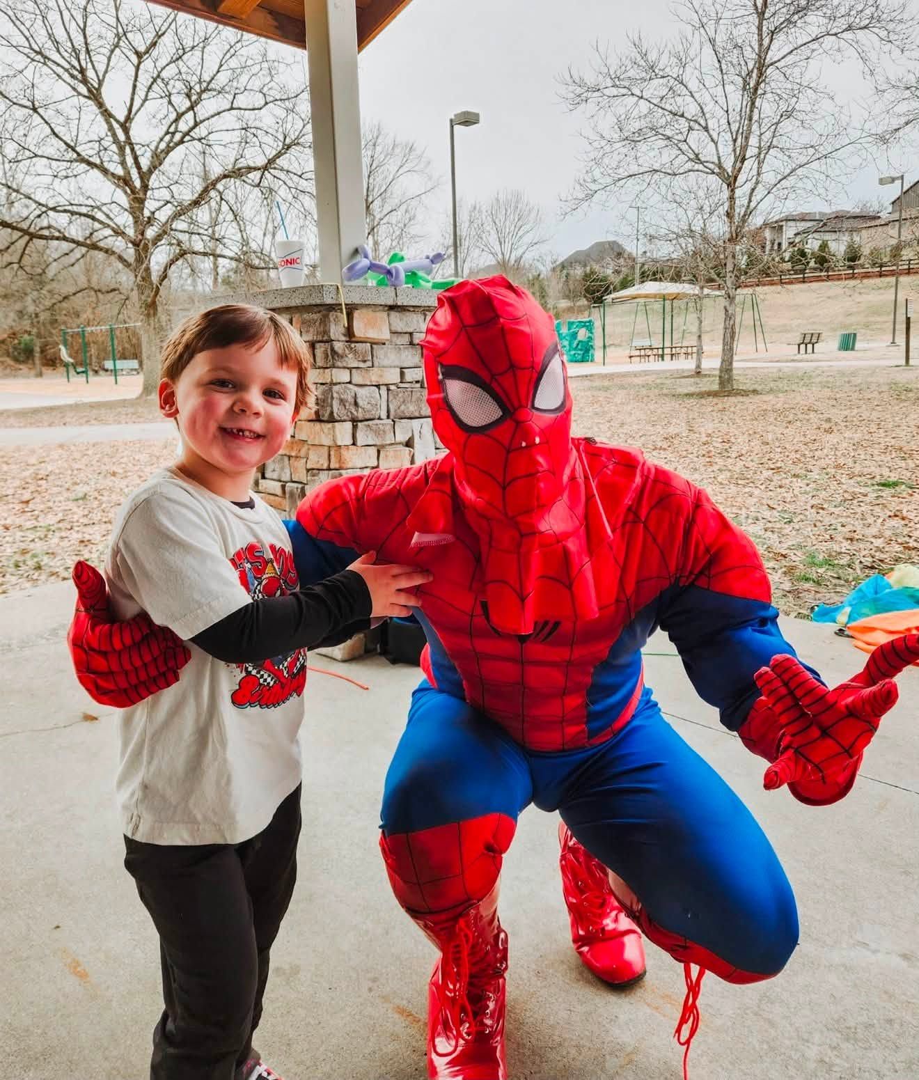 A superhero performer in a red and blue web-patterned suit kneeling beside a beaming young guest at a park pavilion