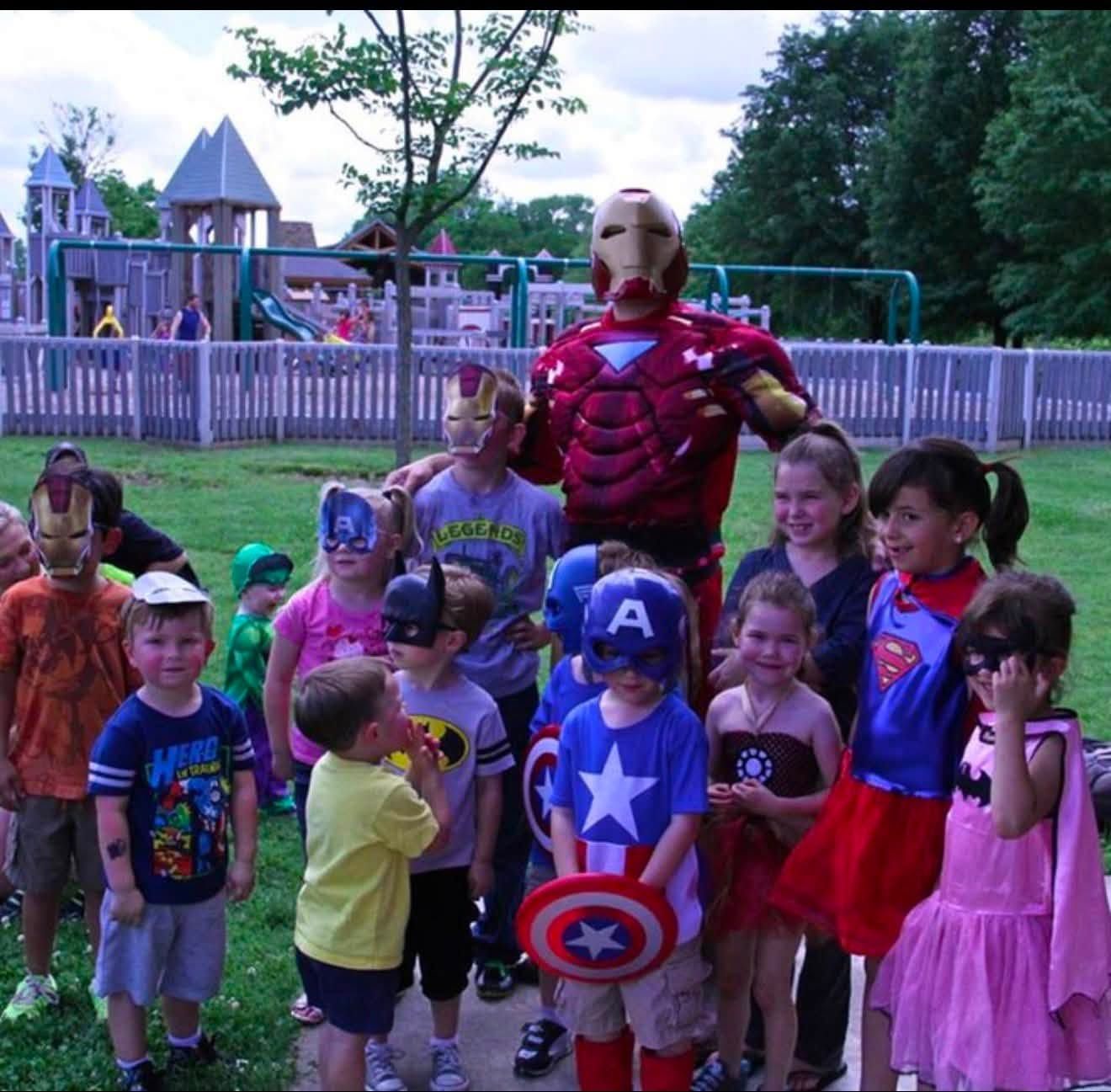 An armored superhero performer surrounded by children in superhero masks, capes and costumes at a playground party