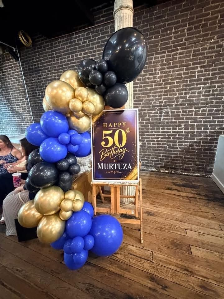 A black, gold and blue balloon garland beside a personalized 50th birthday welcome sign on an easel against a brick wall