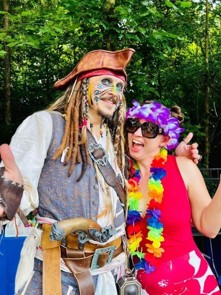 A pirate captain character with braided hair and a tricorn hat posing with a laughing guest wearing flower leis
