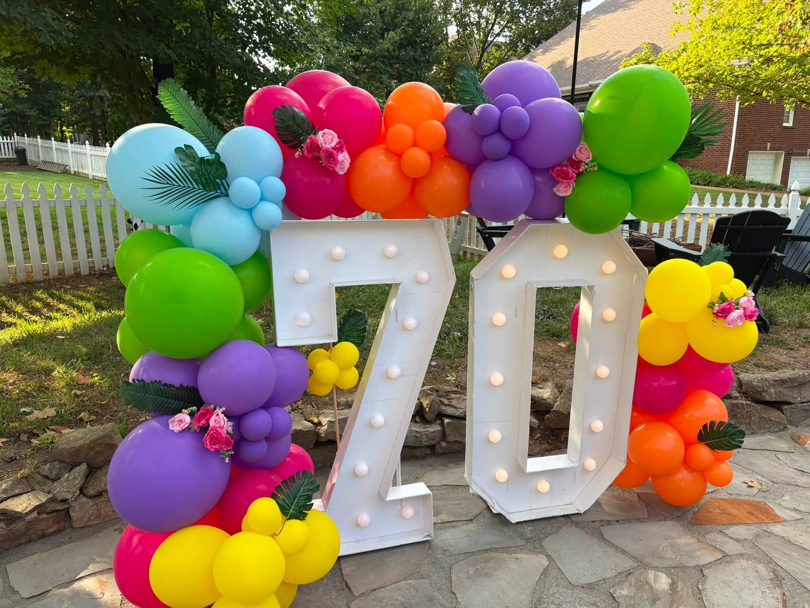 Lit marquee 70 numbers wrapped in a bright balloon garland of pink, orange, purple and green with roses and palm leaves