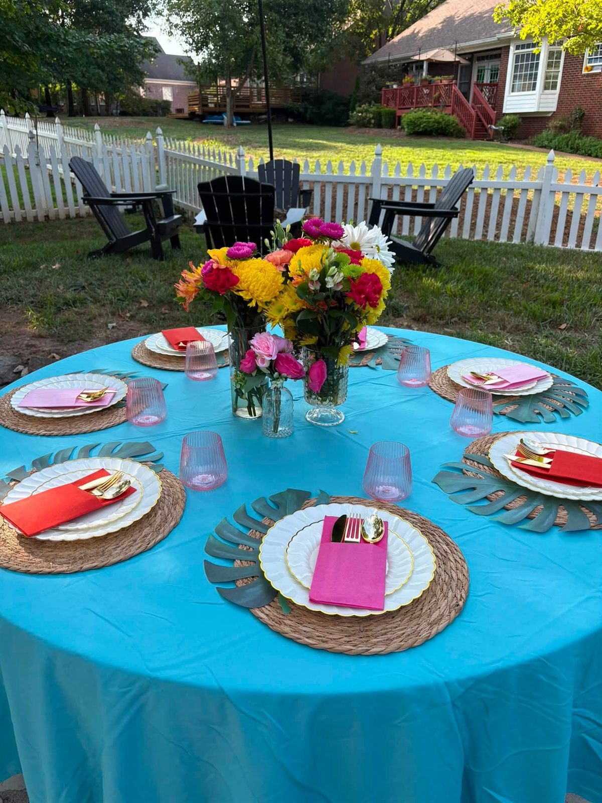 A round party table with turquoise linens, monstera leaf placemats, woven chargers, scalloped plates and bright pink and orange napkins, centered with colorful garden flowers