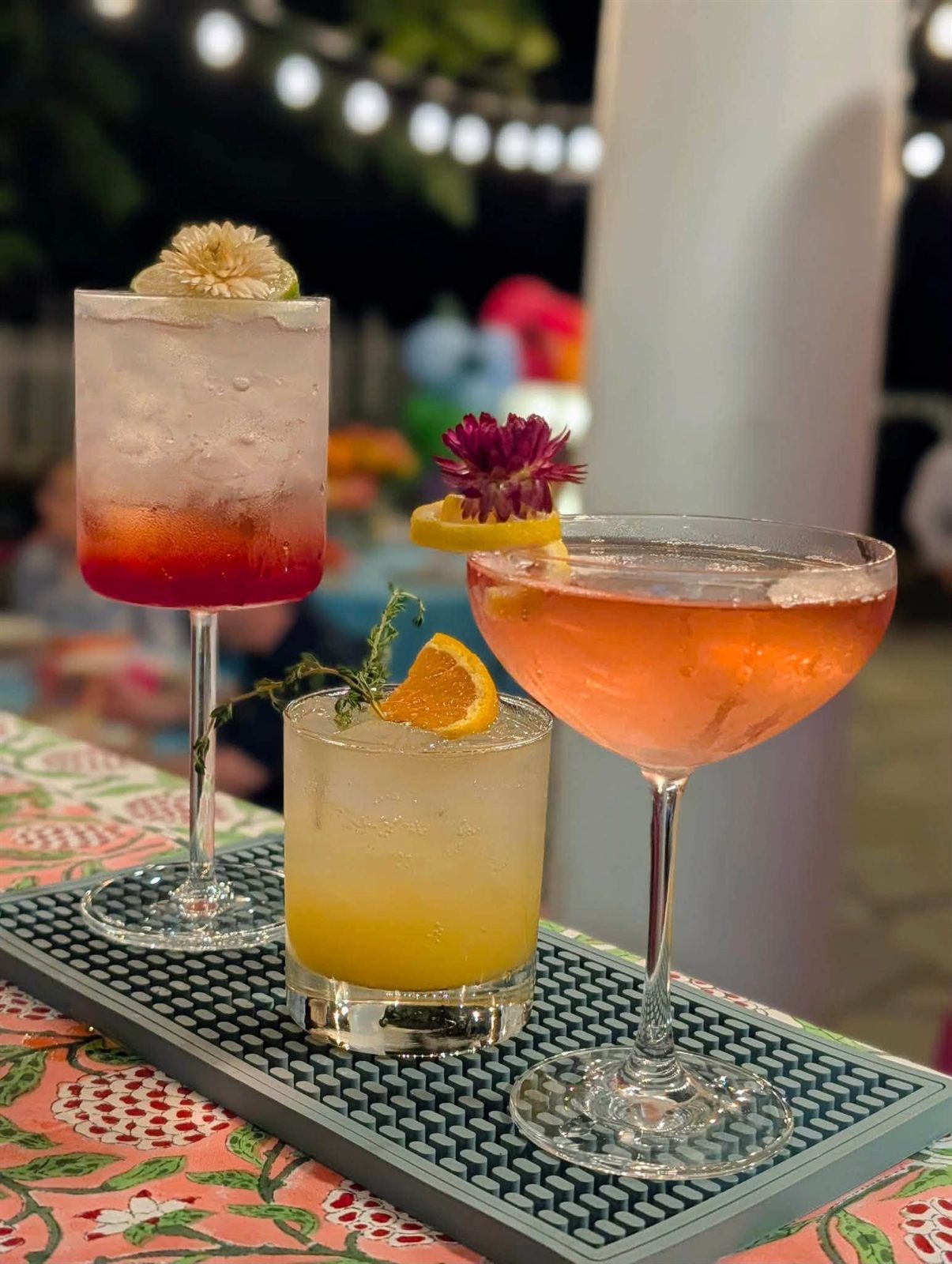 Three garnished signature cocktails in different glasses on a bar mat at night with string lights glowing behind