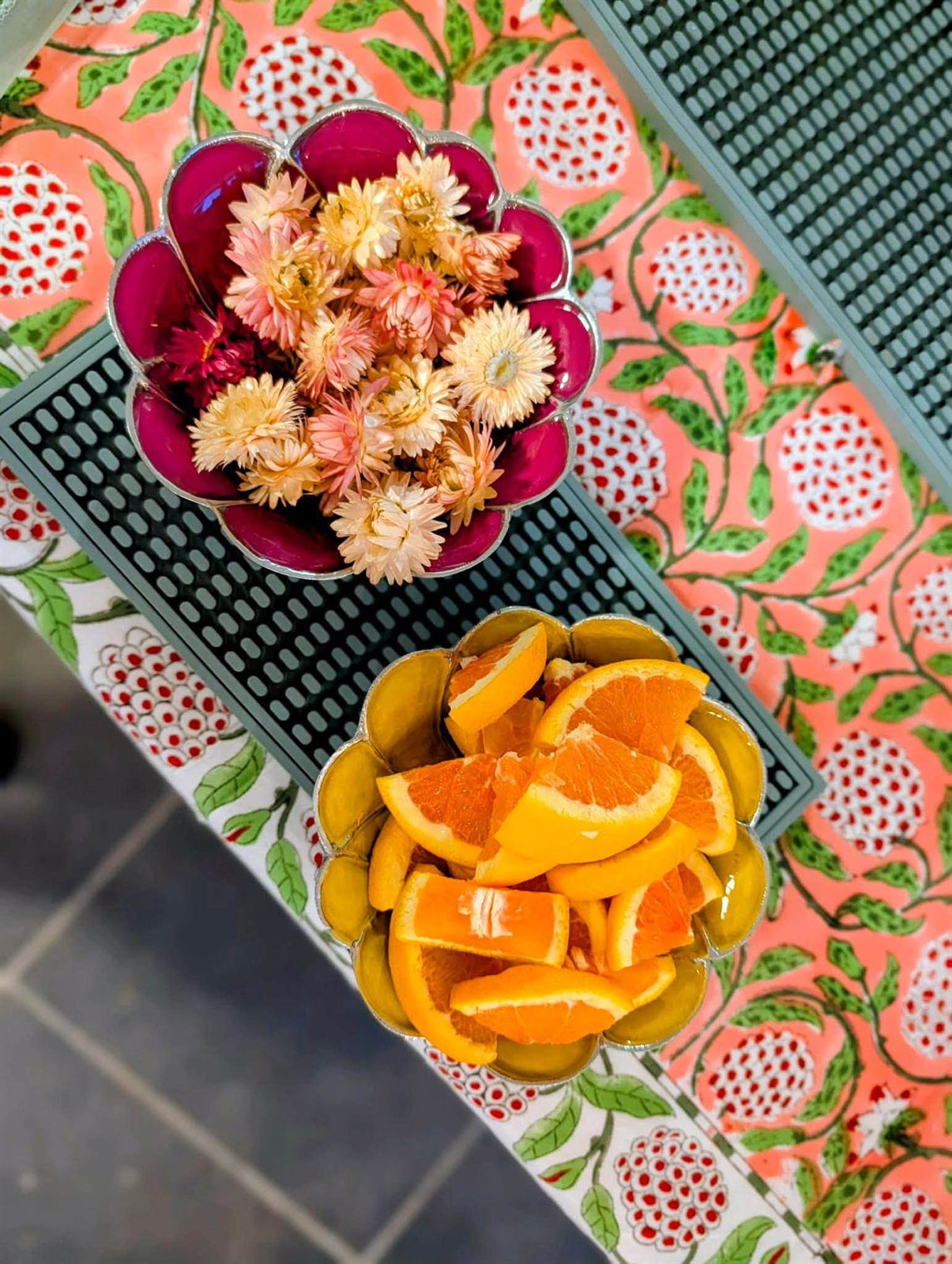 Overhead view of a scalloped bowl of dried flowers and a bowl of fresh orange slices on floral block-print linens